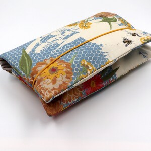 May include: A folded fabric clutch with a floral and bee pattern. The clutch features a blue honeycomb design, orange and red flowers, and a yellow elastic band. The fabric has a cream background with bees and floral accents.