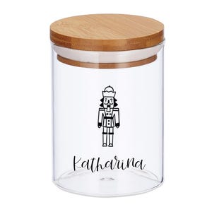 May include: A clear glass jar with a wooden lid. The jar features a black and white illustration of a nutcracker and the name "Katharina" in a script font. The jar is designed for storage.