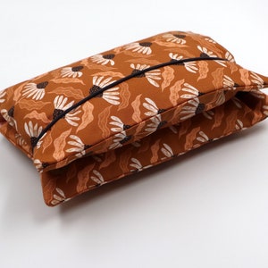 May include: A folded, rectangular pillow with a floral pattern. The pillow features a warm brown background with white and light brown flower and leaf designs. A black trim outlines the pillow's edges.