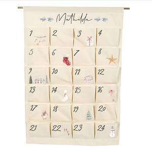 May include: A cream-colored fabric advent calendar hanging from a wooden rod, featuring the name Mathilda at the top. It has twenty-four individual pockets, each numbered one through twenty-four, decorated with small watercolor-style holiday illustrations.