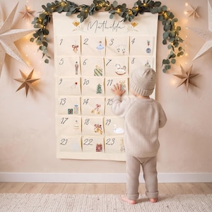 Pode incluir: Um calendário do advento de tecido na cor creme pendurado na parede, com o nome Mathilda, os números de 1 a 23 e várias pequenas ilustrações em aquarela em cada bolso, incluindo uma bailarina, um cavalo de balanço e presentes.