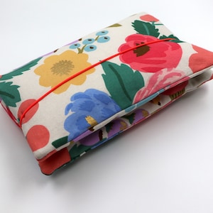 Könnte beinhalten: Eine Clutch mit floralem Muster und rotem Gummibandverschluss. Der Stoff zeigt große, farbenfrohe Blumen in Rosa, Gelb, Blau und Lila auf cremefarbenem Hintergrund. Die Clutch ist gefaltet und zeigt das Innenfutter.