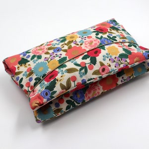 Könnte beinhalten: Eine Clutch aus Stoff mit floralem Muster und einem goldenen Reißverschluss. Die Clutch ist in der Mitte gefaltet und der Stoff ist cremefarben mit rosa, roten, blauen und grünen Blumen.