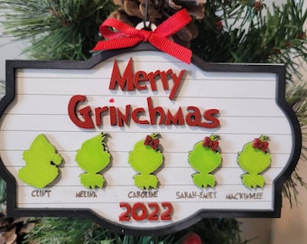 May include: A Christmas ornament with a white background and black border. The words "Merry Grinchmas" are in red, with green Grinch faces below. Names are written under each Grinch face. A red bow and ribbon are at the top.