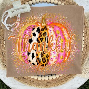 May include: A tan t-shirt featuring a large graphic of a pumpkin with varied patterns including leopard print, cow print, floral, and smiley faces. The word "thankful" is written in orange cursive script across the center of the pumpkin.