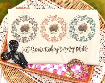 May include: A beige t-shirt featuring three circular designs, each containing a turkey illustration surrounded by a floral border in brown, orange, and green. Below the designs, the text reads, "Put some turkey on my plate." A black hair clip and a stack of beaded bracelets are placed nearby.