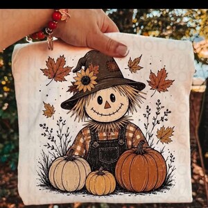 May include: A white t-shirt featuring a graphic illustration of a scarecrow wearing a hat adorned with a sunflower. The scarecrow is dressed in plaid and overalls, surrounded by three pumpkins and falling autumn leaves on a white background.