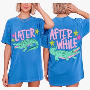 May include: Blue t-shirts with a graphic of an alligator and the text "LATER" on the front and "AFTER WHILE" on the back. The alligator is green with pink accents. The text is pink with yellow and pink star accents.