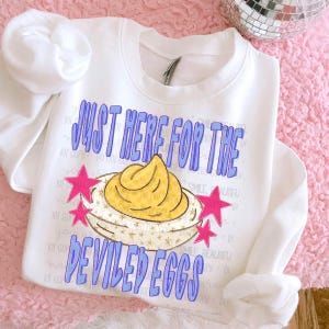 May include: A white long-sleeve sweatshirt featuring a graphic of a deviled egg with a yellow yolk and a white base. The text reads "JUST HERE FOR THE DEVILED EGGS" in purple letters, accented by four bright pink stars on the sides.