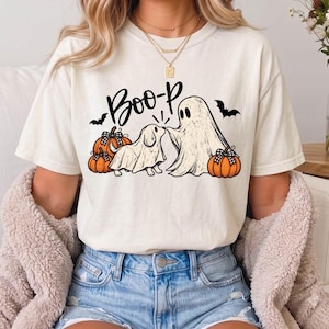 May include: A cream-colored short-sleeve t-shirt featuring a graphic print of a dog and a ghost wearing sheets, with the text "Boo-p" above them. The design includes two orange pumpkins with black and white checkered bows and two black bats.