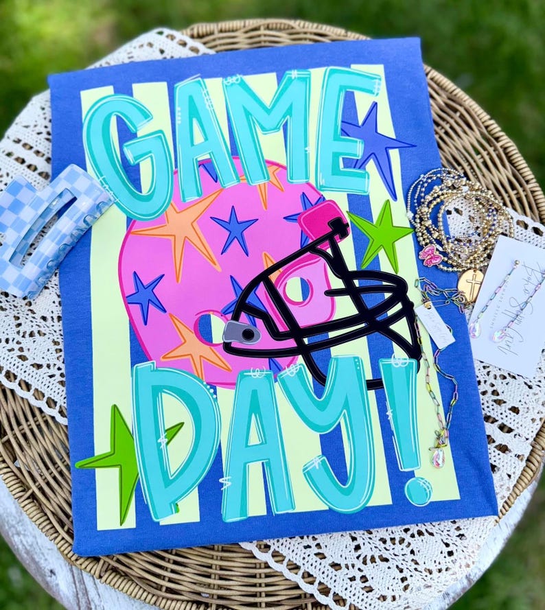May include: A blue t-shirt featuring the text GAME DAY in teal with a pink football helmet graphic decorated with blue, orange, and green stars. The shirt is displayed with a blue and white checkered hair clip and several gold-toned beaded bracelets.