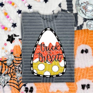 May include: A dark gray striped t-shirt featuring a large, central graphic of a candy corn shape. The graphic is divided into white, orange, and yellow sections with the black cursive text "Trick or treat" printed across the center.