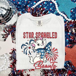 May include: White t-shirt with the words "STAR SPANGLED BEAUTY" in red script. A cartoon cat with a blue bow and fireworks graphic is featured on the front. The shirt is a Comfort Colors brand.
