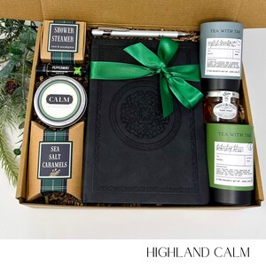May include: A gift box with a black journal tied with a green ribbon, a pen, tea, honey, a shower steamer, lip balm, sea salt caramels, and a tin labeled "CALM." The box is labeled "HIGHLAND CALM."