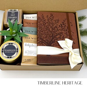 May include: A gift box containing a brown leather journal with a tree design, a gold-colored candle, a small succulent, tea sachets, and a box of sea salt caramels. The journal is tied with a cream-colored ribbon.