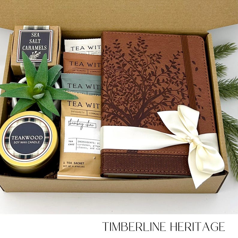 May include: A gift box containing a brown leather journal with a tree design, a gold-colored candle, a small succulent, tea sachets, and a box of sea salt caramels. The journal is tied with a cream-colored ribbon.