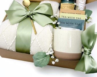 May include: A gift box filled with self-care items, including a white blanket with a green ribbon, a speckled mug, tea bags, a candle, and a small bottle of lotion. The box is decorated with green leaves and white flowers. The text "FRESH NEW DAY" is printed on the bottom of the box.