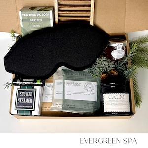 May include: An open gift box filled with spa products. Includes a black eye mask, tea tree oil soap, tea bags, a candle, honey, and a shower steamer. The box is labeled "Evergreen Spa".