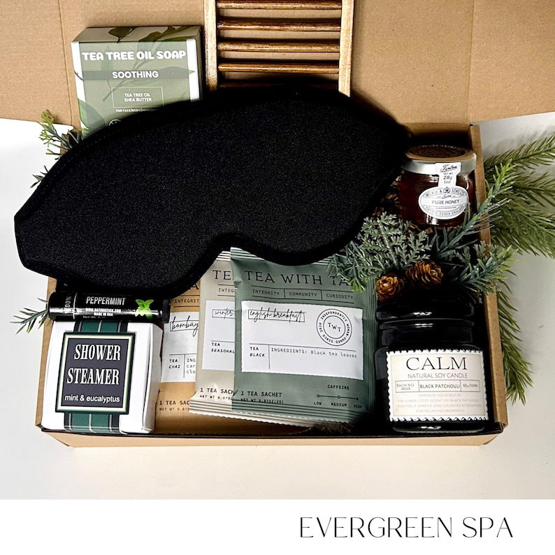 May include: An open gift box filled with spa products. Includes a black eye mask, tea tree oil soap, tea bags, a candle, honey, and a shower steamer. The box is labeled "Evergreen Spa".