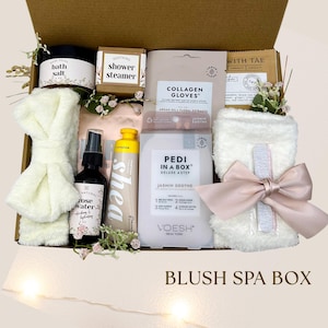May include: A spa gift box filled with self-care products. The box includes bath salt, a shower steamer, rose water, shea lotion, collagen gloves, a foot care kit, a headband, and a washcloth tied with a pink bow. The box is labeled "Blush Spa Box."