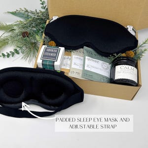 May include: A gift box containing a black padded sleep eye mask with an adjustable strap, a shower steamer, tea sachets, and a jar of natural soy candle labeled "CALM". The box is decorated with greenery.