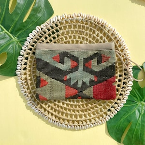 May include: A small, rectangular clutch purse with a woven fabric exterior. The fabric has a geometric pattern in shades of red, black, and green. The purse has a beige fabric lining and a brown leather strap.