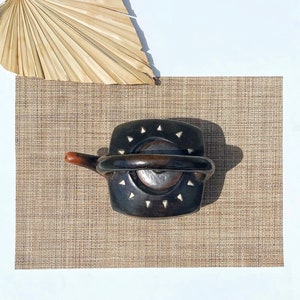 May include: A black ceramic teapot with a wooden handle and white accents. The teapot is sitting on a brown woven placemat.