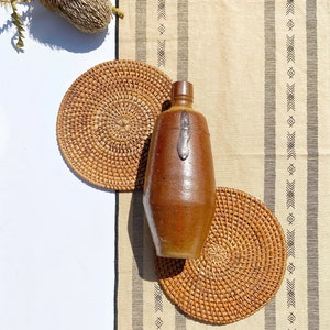 Op de afbeelding: Een bruine keramische fles met een metalen deksel staat op twee geweven rotan-onderzetters. De onderzetters liggen op een beige stof met zwarte geometrische patronen.