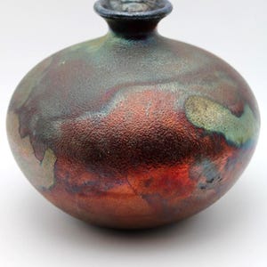May include: A handcrafted ceramic vase with a rounded body and a small, cylindrical neck. The vase features a textured surface with a metallic finish, showcasing hues of red, brown, and green. The vase is approximately 6 inches tall.