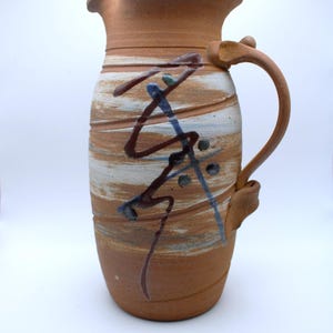 Può includere: Brocca in ceramica fatta a mano con un design marrone, bianco e blu. La brocca ha un manico curvo e un beccuccio per versare. Il corpo della brocca è decorato con linee orizzontali e pennellate astratte.