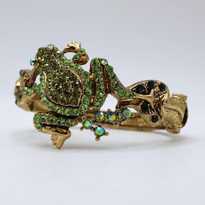 Puede incluir: Un brazalete rígido articulado de metal en tono dorado con un diseño de rana prominente. La rana está incrustada con numerosas piedras de estrás pequeñas de color verde vibrante, y sus patas tienen cristales circulares iridiscentes.