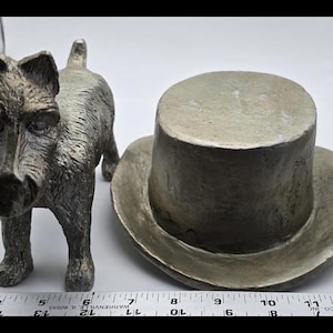 May include: A silver-coloured metal sculpture of a dog wearing a silver-coloured top hat. The dog is standing on a white surface.