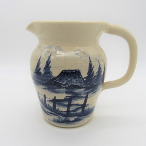 以下が含まれることがあります： クリーム色のセラミックピッチャー。青と白の手描きで、山、木々、フェンスが描かれています。ピッチャーは丸みを帯びたボディとハンドルが付いています。