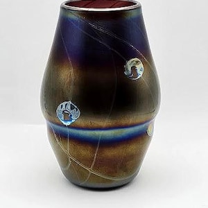 Pode incluir: Vaso de vidro artesanal com um acabamento iridescente único. O vaso transita de tons castanhos escuros para azul e roxo, com detalhes decorativos em vidro transparente. Possui uma base larga e um gargalo mais estreito, adequado para arranjos florais.