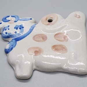 Puede incluir: Adorno de cerámica blanca con forma de vaca con detalles pintados en azul y rosa. La vaca tiene un agujero en la parte superior para colgar.