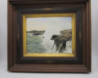 Op de afbeelding: Vierkant ingelijst schilderij van een kustlandschap met golven. Het kunstwerk heeft een bruine houten lijst met gouden accenten. De schildering toont een rotsachtige kustlijn met een woelige zee en een lichtblauwe lucht.