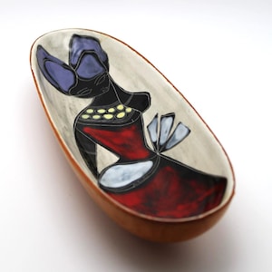 May include: Oval ceramic dish with a hand-painted design. The interior features a stylized figure in black, red, and white, with a purple head covering and yellow accents. The dish has a brown rim and a light gray background.