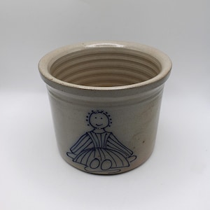 Puede incluir: Un tarro de gres con un exterior gris claro y una ilustración azul de una persona con un vestido. El recipiente cilíndrico tiene una abertura ancha y un interior texturizado. El diseño es simple y rústico.