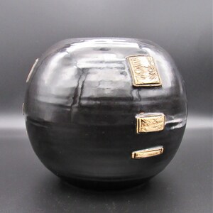 Può includere: Un vaso in ceramica nero con forma rotonda e dettagli dorati. Il vaso ha una finitura lucida ed è decorato con forme geometriche.