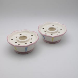 May include: Two white ceramic flower frog vases with pink and blue accents. Each vase has a scalloped rim and a central hole with multiple smaller holes around it.