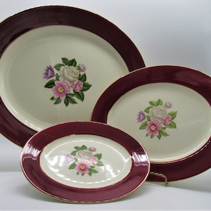 May include: A set of three oval serving platters. Each platter features a cream-coloured centre with a floral design of pink, purple, and white flowers. The rims are a deep burgundy with a gold trim.
