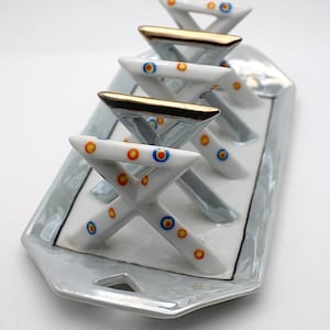 Pode incluir: Bandeja de servir decorativa em cerâmica com cinco suportes em forma de X. Os suportes são brancos com pontos laranja, azuis e amarelos. A bandeja tem um acabamento perolado e detalhes dourados.