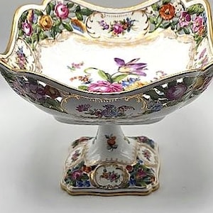 May include: A white porcelain footed serving dish with a scalloped rim and gold trim. The dish is decorated with colorful floral patterns, including roses and other flowers. The base is square and also decorated with flowers.