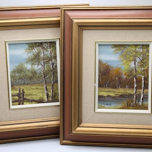 May include: Two framed landscape paintings. Each features a serene outdoor scene with trees, a grassy field, and a blue sky. The frames are brown with gold inner edges, and the paintings are displayed on a neutral linen-like background.