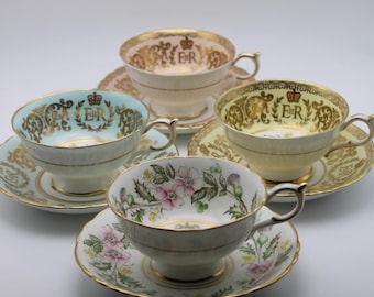 Könnte beinhalten: Vier Vintage-Teetassen und Untertassen in Pastellfarben, darunter Rosa, Gelb und Blau. Jede Tasse hat goldene Verzierungen und verzierte Designs, einige tragen die Initialen "ER" und eine Krone. Eine Tasse zeigt ein Blumenmuster.