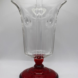 May include: A clear glass vase with a flared top and a ruby red base. The vase has a decorative design near the top and a series of vertical lines. The base is round and has a stem that connects to the clear glass portion.
