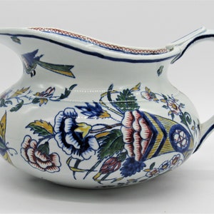 Könnte beinhalten: Weißer Keramikkrug mit breitem Körper und gebogenem Henkel. Der Krug ist mit farbenfrohen Blumen- und Blattmustern in Rot, Blau, Gelb und Grün verziert. Der Rand und der Henkel sind blau umrandet.