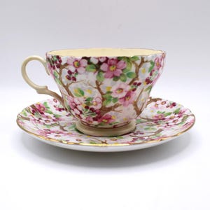 May include: A floral teacup and saucer set. The cup and saucer are decorated with pink flowers, green leaves, and brown branches. The rim of the cup and saucer is trimmed with gold. The handle of the cup is a light beige colour.