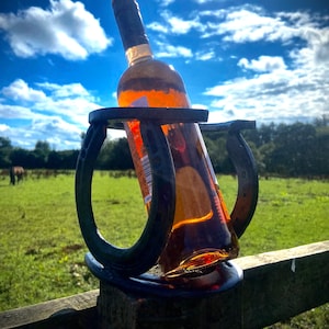 May include: A bottle of wine held in a horseshoe bottle holder. The bottle holder is made of metal and is attached to a wooden fence post. The bottle is a dark amber colour and the wine is visible through the glass. The background is a blue sky with white clouds.