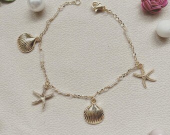 Op de afbeelding: Een gouden kettingarmband met witte kralen en bedels. De bedels omvatten twee schelpen en twee zeesterren.